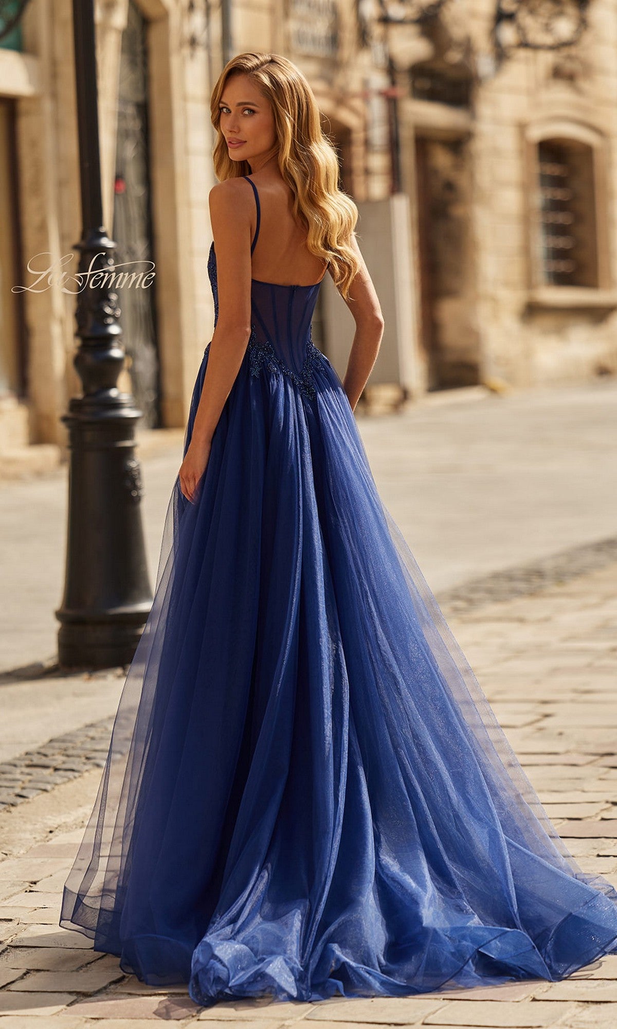 A woman wearing the La Femme 33436 long prom dress—sleeveless with thin straps and a high leg slit—stands outdoors on a cobblestone street, glancing back over her shoulder.